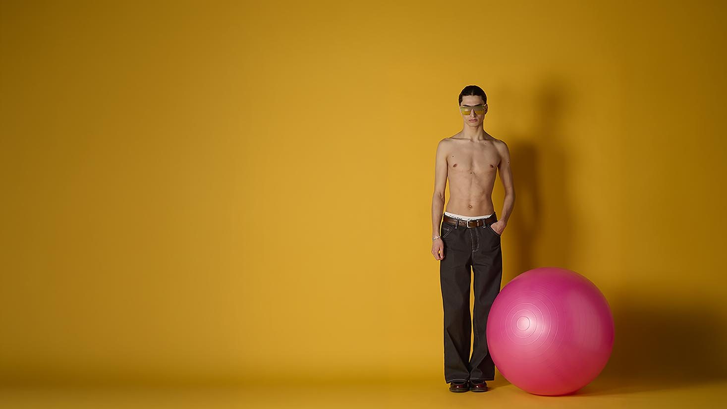 Model in black jeans and tinted sunglasses stands beside a pink exercise ball against a mustard yellow wall